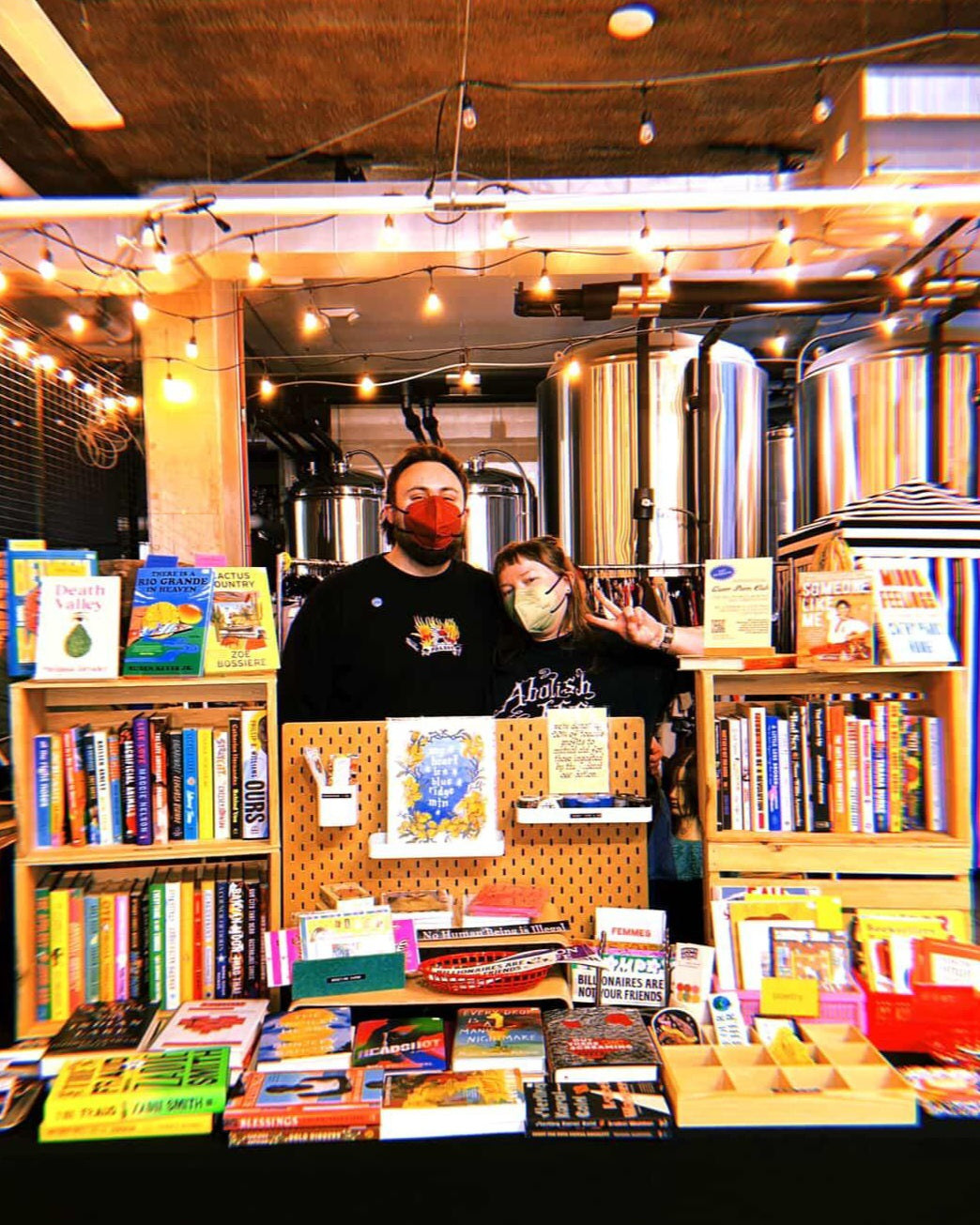 Two masked people (Beck and Ty) standing behind a display table filled with books in a brewery (decorative)